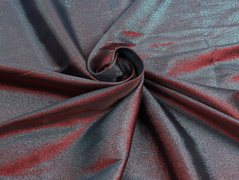 May include: Close-up of a metallic fabric with a blue and red iridescent sheen. The fabric is folded and draped, creating a soft, flowing texture.