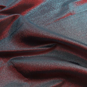 May include: Close-up of a shimmering fabric with a blue and red iridescent sheen. The fabric is draped and folded, creating soft, flowing lines.