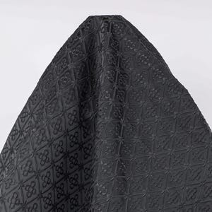 K&ouml;nnte beinhalten: Ein dunkelgrauer Stoff mit einem sich wiederholenden gepr&auml;gten Muster aus stilisierten floralen Motiven. Der Stoff ist &uuml;ber eine unsichtbare Form drapiert und bildet eine dreieckige Form. Die Textur scheint weich und geschmeidig zu sein.