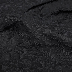 Puede incluir: Primer plano de una tela negra con un patrón floral en relieve. La tela tiene una superficie texturizada con un diseño repetido de flores y hojas estilizadas. El color oscuro y los detalles intrincados crean una apariencia lujosa.