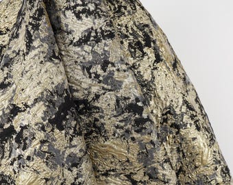 May include: Close-up of a textured fabric with a pattern of black, gray, and gold. The fabric is folded, revealing the intricate details of the design. The material has a shimmering, metallic appearance.