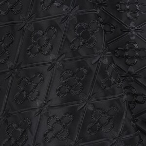K&ouml;nnte beinhalten: Nahaufnahme eines schwarzen Stoffes mit einem sich wiederholenden Muster. Das Muster besteht aus Rauten mit bl&uuml;tenartigen Designs in der Mitte und an den Ecken. Der Stoff hat einen dezenten Glanz, der ihm ein luxuri&ouml;ses Aussehen verleiht.