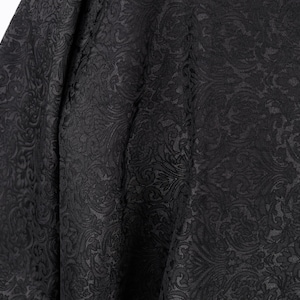 Op de afbeelding: Close-up van zwarte stof met een gedetailleerd, in reliëf aangebracht bloemmotief. De stof heeft een subtiele glans en het ontwerp is consistent over het oppervlak. De plooien in de stof creëren schaduwen die de textuur benadrukken.