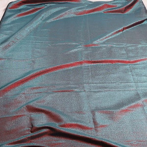 May include: A teal and burgundy iridescent fabric with a subtle shimmer. The fabric has a woven texture and a slightly crinkled appearance.