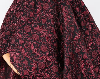 Op de afbeelding: Zwarte stof met een herhaald patroon van rode bloemen- en krulwerkontwerpen. De stof heeft een getextureerd uiterlijk en is waarschijnlijk geschikt voor kleding of woondecoratie.