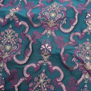 May include: Teal fabric featuring a repeating pattern of ornate floral and scroll designs in purple and gold. The fabric has a slight sheen, suggesting a luxurious material.