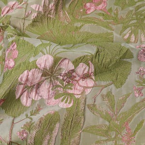Könnte beinhalten: Nahaufnahme eines Stoffes mit floralem Muster. Der Stoff zeigt große rosa Blüten und grüne Blätter auf hellgrünem Hintergrund. Das Design hat ein strukturiertes, geprägtes Aussehen, das Tiefe und visuelles Interesse erzeugt. Der Stoff wird wahrscheinlich für Kleidung oder Heimdekoration verwendet.