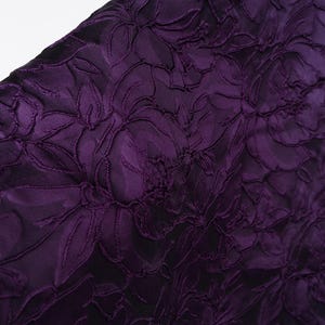Pu&ograve; includere: Primo piano di un tessuto viola scuro con un motivo floreale. Il design presenta fiori e foglie strutturate in rilievo su uno sfondo pi&ugrave; scuro, creando un effetto dimensionale. Il tessuto ha un aspetto lussuoso ed elegante.