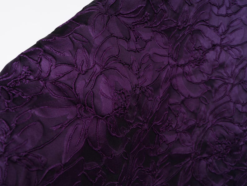 Pu&ograve; includere: Primo piano di un tessuto viola scuro con un motivo floreale. Il design presenta fiori e foglie strutturate in rilievo su uno sfondo pi&ugrave; scuro, creando un effetto dimensionale. Il tessuto ha un aspetto lussuoso ed elegante.