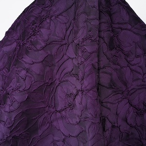 Pu&ograve; includere: Primo piano di un tessuto viola scuro con un motivo floreale. Il tessuto presenta un design strutturato e in rilievo di grandi fiori e foglie. Il colore &egrave; una ricca tonalit&agrave; scura di viola.