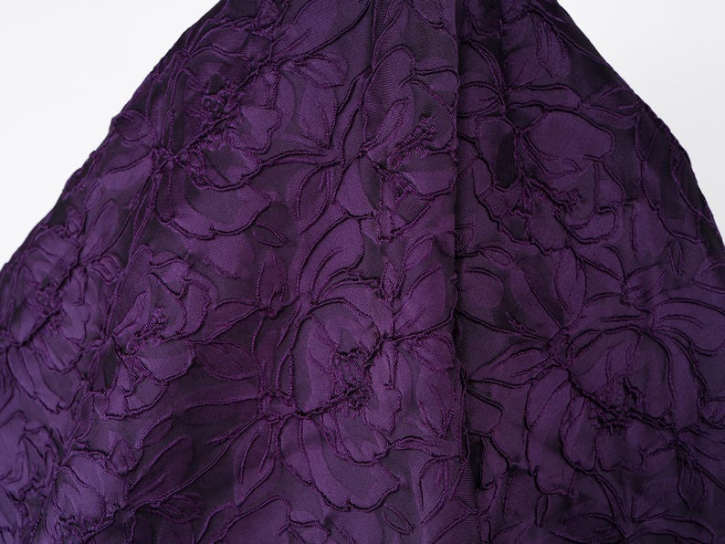 Pu&ograve; includere: Primo piano di un tessuto viola scuro con un motivo floreale. Il tessuto presenta un design strutturato e in rilievo di grandi fiori e foglie. Il colore &egrave; una ricca tonalit&agrave; scura di viola.