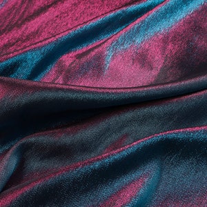 May include: Close-up of a blue and pink iridescent fabric with a textured surface. The fabric is folded and draped, showing its shimmery and reflective qualities.