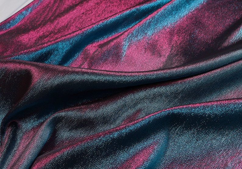 May include: Close-up of a blue and pink iridescent fabric with a textured surface. The fabric is folded and draped, showing its shimmery and reflective qualities.