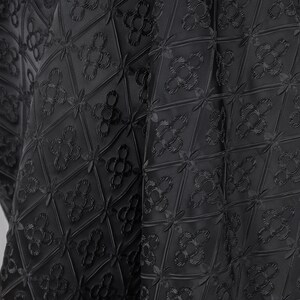 K&ouml;nnte beinhalten: Schwarzer Stoff mit einem sich wiederholenden geometrischen Muster aus Rauten und floralen Motiven. Der Stoff scheint strukturiert zu sein, mit erhabenen Details, die ein subtiles Muster erzeugen. Der Gesamteffekt ist ein dunkles, elegantes Design.