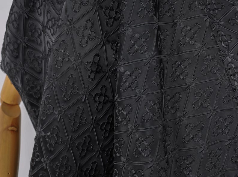 K&ouml;nnte beinhalten: Schwarzer Stoff mit einem sich wiederholenden geometrischen Muster aus Rauten und floralen Motiven. Der Stoff scheint strukturiert zu sein, mit erhabenen Details, die ein subtiles Muster erzeugen. Der Gesamteffekt ist ein dunkles, elegantes Design.