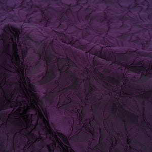 Pu&ograve; includere: Primo piano di un tessuto viola scuro con un motivo floreale. Il tessuto presenta un design strutturato e in rilievo, che crea un effetto tridimensionale. Le pieghe e le ombre aggiungono profondit&agrave; al colore ricco e scuro.