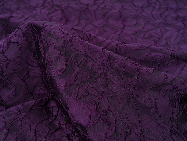 Pu&ograve; includere: Primo piano di un tessuto viola scuro con un motivo floreale. Il tessuto presenta un design strutturato e in rilievo, che crea un effetto tridimensionale. Le pieghe e le ombre aggiungono profondit&agrave; al colore ricco e scuro.