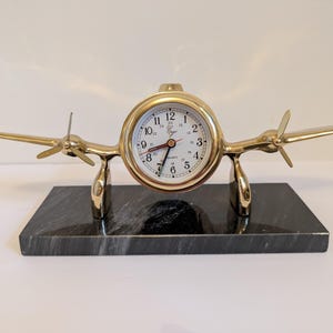 May include: A brass airplane-shaped clock with a white clock face and black numerals. The clock has a quartz movement and is mounted on a black marble base. The clock's wings and propellers are made of brass.