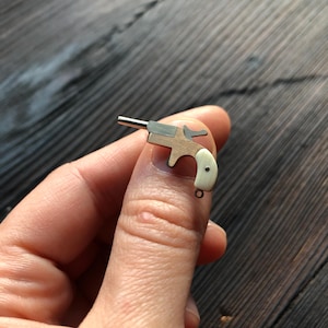 May include: A miniature pistol charm featuring a metallic barrel and frame with a smooth, off-white handle grip. The small firearm replica includes a tiny loop at the base of the handle for attachment, showcasing a multi-material construction.