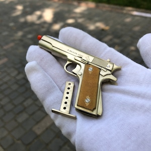 Könnte beinhalten: Eine silberne Spielzeug-Nachbildung einer US-Army 1911 Pistole mit braunem Kunststoffgriff und Magazin. Die Spielzeugpistole befindet sich in einem weißen Handschuh.