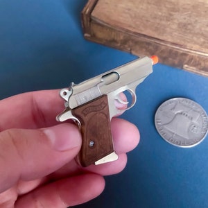 Könnte beinhalten: Eine kleine, silberfarbene Pistole mit braunem Griff, mit der Aufschrift "WALTHER". Der Lauf hat eine orangefarbene Spitze. Neben der Pistole ist eine Silbermünze zu sehen. Die Pistole wird in einer Hand gehalten.