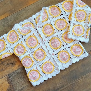 Puede incluir: Una chaqueta de punto de ganchillo blanca con un diseño de limón rosa y amarillo. La chaqueta tiene cierre de botón y mangas largas.