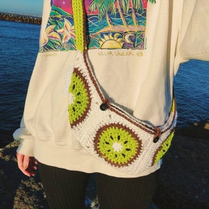 Puede incluir: Un bolso bandolera de crochet blanco con un diseño de kiwi. El bolso tiene una correa marrón y dos botones negros. El bolso lo lleva una persona que lleva una sudadera blanca con un estampado gráfico colorido.