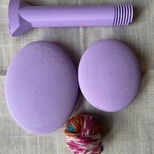 May include: A set of three purple 3D printed weaving tools, including two circular loom bases and a shuttle. A ball of yarn in shades of brown, pink, and teal is also included.