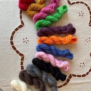 May include: A collection of 12 skeins of yarn in various colours, including red, orange, yellow, green, blue, purple, pink, brown, grey, black, and white. The yarn is arranged in a rainbow pattern on a white tablecloth.