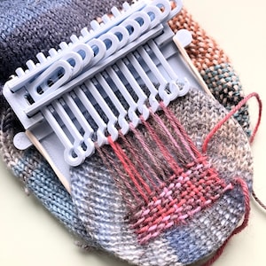 May include: A light blue weaving loom with a partially woven piece of fabric. The fabric is a mix of grey, blue, and red yarn. The loom is made of plastic and has a series of pegs to hold the yarn in place. The project is in progress.