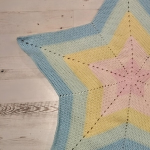 May include: A pastel-coloured, star-shaped crocheted blanket. The blanket has a gradient design, shifting from light blue to yellow and pink in the centre. The handmade blanket has a soft, textured appearance, suitable for a baby or child.