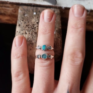 Op de afbeelding: Twee zilveren ringen, elk met een kleine, ovale blauwgroene steen. De ringen hebben een uniek ontwerp, met kleine handen die de steen omarmen. De ringen worden op een vinger getoond.