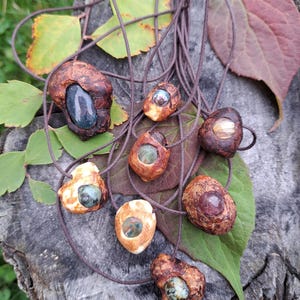 Puede incluir: Una colección de collares hechos a mano con colgantes de madera natural con piedras preciosas incrustadas. Los colgantes varían en forma y tamaño, mostrando una gama de colores desde los tonos naturales de la madera hasta los tonos de las piedras preciosas. Cada collar tiene un cordón marrón.
