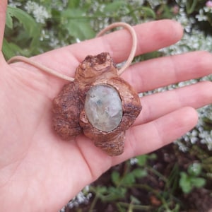 Op de afbeelding: Een handgemaakte hangerketting met een natuurlijke, getextureerde houten basis. De hanger bevat een lichtgroene, ovaalvormige steen. De ketting heeft een lichtbruin koord. De achtergrond is vervaagd met groen gebladerte.