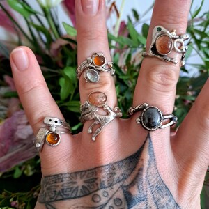 Könnte beinhalten: Eine Hand trägt fünf Silberringe mit verschiedenen farbigen Edelsteinen. Die Ringe haben verschiedene Designs, darunter eine Schlange, einen Ast und ein einfaches Band. Die Edelsteine sind orange, klar und schwarz.