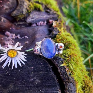 Könnte beinhalten: Ein silberner Ring mit einem großen ovalen blauen Edelstein, umgeben von kleineren silbernen Perlen und einem einzigen orangefarbenen Edelstein. Der Ring liegt auf einem Stück Baumrinde mit Moos darauf.