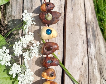 Puede incluir: Un collar hecho a mano con cuentas de madera de forma irregular, en tonos marrones y beige. Cada cuenta está adornada con una piedra o gema de diferente color, ensartada en un cordón marrón. El collar se exhibe sobre una superficie de madera.