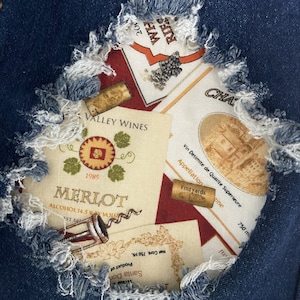 May include: A close-up of ripped denim fabric with a colourful fabric patch underneath. The patch features a collage of wine bottle labels with text that reads "Valley Wines", "Merlot", "1989", "Alcohol 14.5%", "Santa Don", "Product of", "Net Cont. 750 ML", "Vin Delimite de Qualite Superieure", "Appellation", "Vineyards", "750 ml", and "Cha".