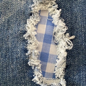 以下が含まれることがあります： 青いジーンズが破れて、その破れ口から青と白のギンガムチェックの生地が見える、クローズアップ写真です。