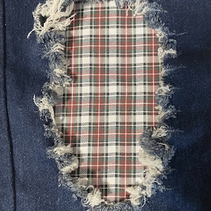 Peut inclure: Gros plan sur un tissu denim délavé avec un patch à carreaux. Le denim bleu présente une section déchirée avec des bords effilochés, révélant un motif à carreaux rouge, vert et blanc en dessous. L'effet général est un look vintage et usé.