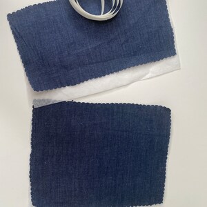 Pu&ograve; includere: Due pezzi di tessuto blu scuro con bordi a festoni, ciascuno posto su un foglio di carta bianca. Un rotolo di nastro adesivo bianco &egrave; posto sul foglio di carta superiore.