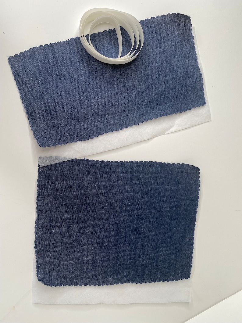 Pu&ograve; includere: Due pezzi di tessuto blu scuro con bordi a festoni, ciascuno posto su un foglio di carta bianca. Un rotolo di nastro adesivo bianco &egrave; posto sul foglio di carta superiore.