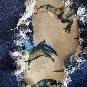 May include: Close-up of distressed denim fabric with a torn section revealing a dinosaur print. The denim is a medium blue with frayed edges. The dinosaur print features various dinosaurs in shades of green, blue, and brown on a beige background.