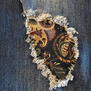 May include: A close-up of a denim fabric with a hole revealing a paisley patterned fabric underneath. The denim has a frayed edge around the hole.