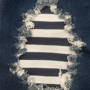 Puede incluir: Un primer plano de un par de pantalones vaqueros azules con un agujero que ha sido remendado con una tela a rayas blancas y azul marino. El agujero está deshilachado y la tela es visible a través del agujero.
