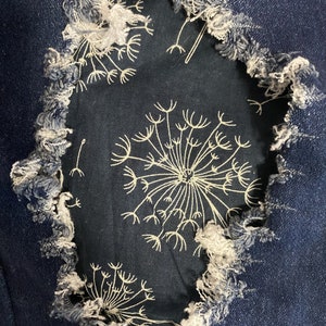 Könnte beinhalten: Nahaufnahme eines dunkelblauen Jeansstoffes mit einem Loch, das einen schwarzen Stoff mit einem weißen Löwenzahnmuster freilegt.