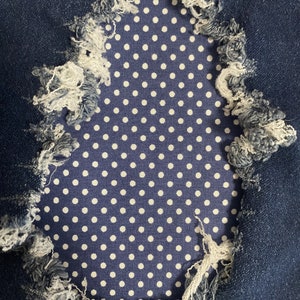 Op de afbeelding: Een close-up van een blauwe denim stof met een gat dat een wit stippenpatroon eronder onthult.