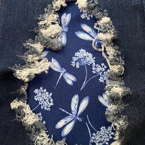 Peut inclure: Gros plan sur une paire de jeans bleus avec un trou déchiré révélant un patch de tissu bleu avec un motif de libellule et de fleurs blanches.