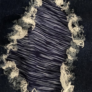 Puede incluir: Primer plano de tela vaquera desgastada con un agujero grande e irregular. Los bordes del agujero están deshilachados con hilos blancos y azules. El interior revela una tela a rayas azules y blancas.