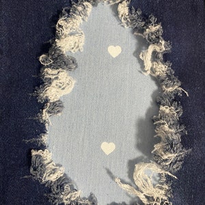 May include: A close-up of a pair of blue denim jeans with a distressed hole revealing a light blue fabric underneath. Two white heart-shaped patches are visible inside the hole.